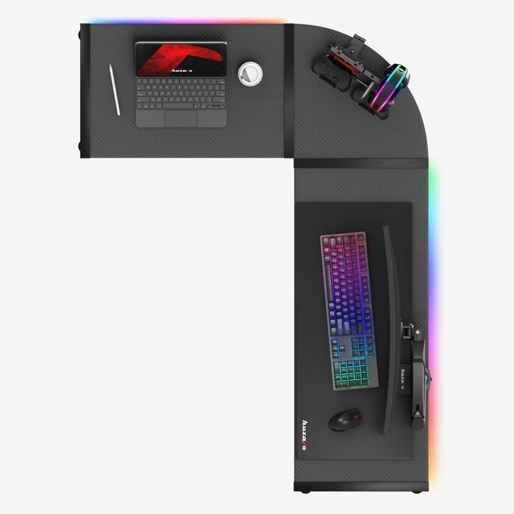 Pogled odozgo na Huzaro Hero 6.0 L-shape RGB gaming sto: tastatura sa višebojnim osvetljenjem, miš, zakrivljeni monitor, laptop, šolja i nosač kontrolera/slušalica na tamnoj karbon površini sa svetlećom RGB trakom